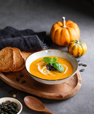 Kremalı balkabağı çorbası, fesleğen ve tohumlar tahtadan bir tahtanın üzerinde koyu bir zemin üzerinde kızarmış ekmek ve taze balkabağı ve bir kaşık. Kırsal tarzda sağlıklı ve samimi sonbahar yemekleri kavramı. Boşluğu kopyala.