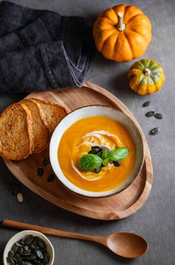 Kremalı balkabağı çorbası, fesleğen ve tohumlar tahtadan bir tahtanın üzerinde koyu bir zemin üzerinde kızarmış ekmek ve taze balkabağı ve bir kaşık. Kırsal tarzda sağlıklı ve samimi sonbahar yemekleri kavramı. Üst görünüm.