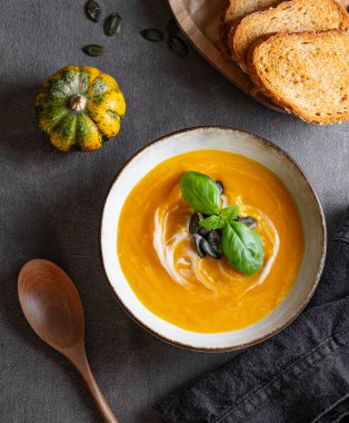 Kremalı, fesleğenli ve tohumlu ev yapımı balkabağı çorbası. Arka planda taze kabaklar, kızarmış ekmek, bitki ve kaşık var. Kırsal tarzda sağlıklı ve samimi sonbahar yemekleri kavramı. Üst görünüm.