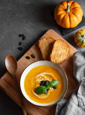 Kremalı balkabağı çorbası, fesleğen ve tohumlar tahtadan bir tahta üzerinde ve koyu arkaplanda kızarmış ekmek ve kaşık. Kırsal tarzda sağlıklı ve samimi sonbahar yemekleri kavramı. Üst görünüm ve kopyalama alanı.