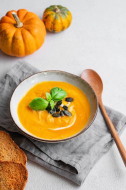 Kremalı, fesleğenli ve tohumlu ev yapımı balkabağı çorbası. Ekmek, peçete, taze balkabağı ve kaşıkla birlikte. Sağlıklı ve samimi sonbahar yemekleri kavramı. Üst görünüm ve kopyalama alanı.