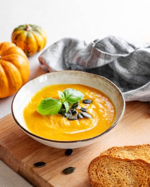 Kremalı, fesleğenli ve tohumlu ev yapımı balkabağı çorbası ahşap bir tahtada ekmek ve peçeteyle birlikte taze balkabaklarıyla birlikte. Sağlıklı ve samimi sonbahar yemekleri kavramı. Boşluğu kopyala.