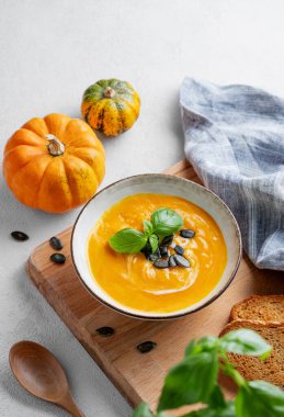 Kremalı balkabağı çorbası, fesleğen ve tohumlar tahta bir tahtanın üzerinde hafif bir zemin üzerinde taze balkabağı ve bitkiyle birlikte. Sağlıklı ve samimi sonbahar yemekleri kavramı. Üst görünüm ve kopyalama alanı.