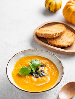 Kremalı balkabağı çorbası, fesleğen ve tohumlar arka planda ekmek ve kaşıkla. Sağlıklı ve samimi sonbahar yemekleri kavramı. Üst görünüm ve kopyalama alanı.