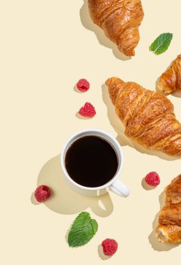 Düz kahve fincanı, taze kruvasanlar, ahududu ve sarı arka planda gölgeli nane yaprağı. Yaratıcı tasarım ve sağlıklı yemek ve Fransız kahvaltısı konsepti. Üst görünüm ve kopyalama alanı.