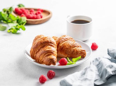 Bir fincan espresso kahve ve taze ahududu ile kruvasan ve sabah ışığı ve peçeteli beyaz desenli bir tabakta nane şekeri. Sağlıklı yemek ve Fransız kahvaltısı kavramı. 