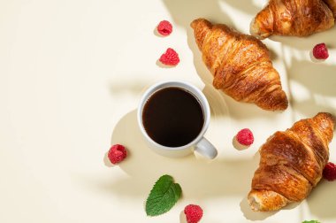 Düz kahve fincanı, taze kruvasanlar, ahududu ve sarı arka planda gölgeli nane yaprağı. Yaratıcı tasarım ve sağlıklı yemek ve Fransız kahvaltısı konsepti. Üst görünüm ve kopyalama alanı.