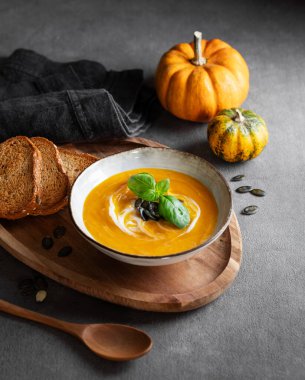 Kremalı balkabağı çorbası, fesleğen ve tohumlar tahtadan bir tahtanın üzerinde, koyu bir zemin üzerinde kızarmış ekmek ve taze balkabağı ve kaşık. Kırsal tarzda sağlıklı ve samimi sonbahar yemekleri kavramı. Boşluğu kopyala.