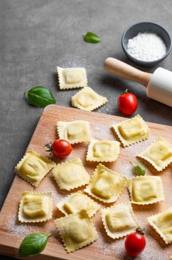 Koyu arka planda ahşap kesim tahtasıyla domates, un, fesleğen ve oklavayla doldurulmuş ev yapımı makarna ravioli. Geleneksel İtalyan yemeği konsepti. Boşluğu kopyala.