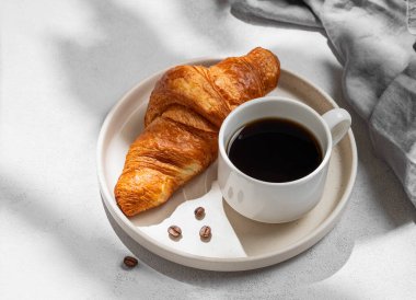 Bir fincan espresso kahve ve beyaz tabakta taze kruvasan gölge ve peçeteyle desenli arka planda. Yaratıcı bir maket ve sağlıklı yemek ve Fransız kahvaltısı konsepti. Üst görünüm ve kopyalama alanı