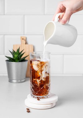 Uzun bardakta soğuk latte kahve. Süt sürahisinden kahve çekirdekleri ve ev bitkisi olan beyaz tuğlalı bir duvara karşı. Yaz modası içki konsepti.