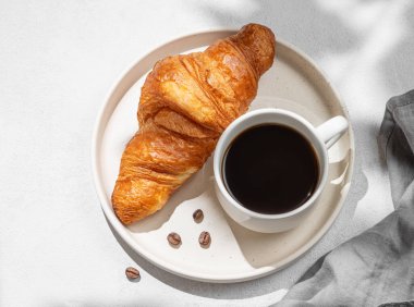 Bir fincan espresso kahve ve beyaz tabakta taze kruvasan gölge ve peçeteyle desenli arka planda. Yaratıcı bir maket ve sağlıklı yemek ve Fransız kahvaltısı konsepti. Üst görünüm ve kopyalama alanı