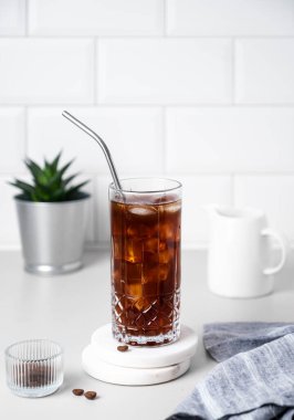 Kahve çekirdekleri, ev bitkisi, süt sürahisi ve peçeteli beyaz tuğlalı arka planda buzlu ve pipetli soğuk bir kahve. Yaz ferahlatıcı modern içki. Ön görünüm.