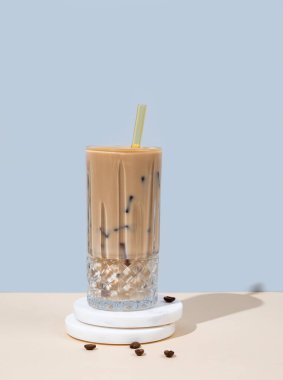 Sütlü büyük bardakta soğuk latte kahve ve kahve çekirdekleri ve gölgeli sarı ve mavi arka planda mermer tezgahta pipet. Yaz modası içeceği. Ön görünüm ve boşluk kopyala.