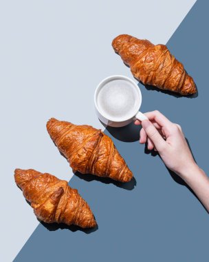 El ele tutuşan kapuçino ya da mavi arka planda taze kruvasan ve gölgeli latte kahve. Kafe reklamları ya da Fransız kahvaltı konsepti için yaratıcı bir model. Üst görünüm ve kopyalama alanı.
