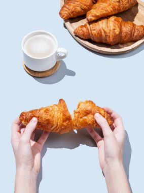 Mavi arka planda bir fincan kapuçino ya da latte kahve ve hamur işiyle taze kruvasan kıran eller. Kafe reklamları ya da Fransız kahvaltı konsepti için yaratıcı bir model. Üst görünüm ve kopyalama alanı.