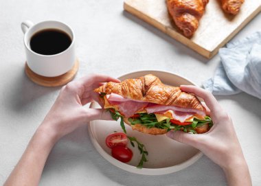 Elleri taze kruvasan sandviçi, jambon, peynir, domates ve roka ile hafif bir arka planda bir fincan kahve ve taze hamur işi ile birlikte. Sağlıklı bir kahvaltı için kavram. Üst görünüm.