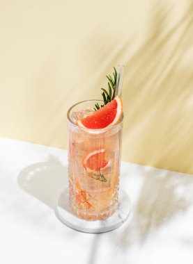 Beyaz sarı arka planda gölgeli bir bardakta greyfurt, biberiye ve buzla mocktail tazeleyici. Yaz turunçgil içeceği ya da kafe reklamları için limonata konsepti. Boşluğu kopyala.