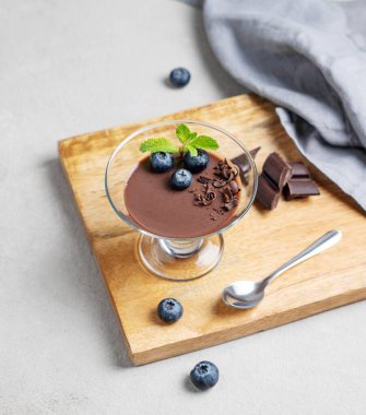 Taze yaban mersinli bir bardakta çikolatalı puding ve arka planda böğürtlen, kaşık ve peçeteli bir tahta üzerinde nane şekeri. Ev yapımı kremalı tatlı ya da panna cotta konsepti. Üst görünüm