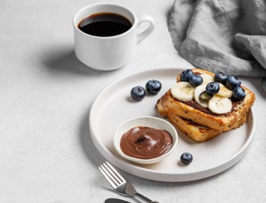 Çikolata kremalı tost ekmeği, yaban mersini ve muz bir tabak hafif arka planda bir fincan espresso kahvesiyle birlikte. Lezzetli ve sağlıklı bir Fransız kahvaltısı kavramı. Boşluğu kopyala.