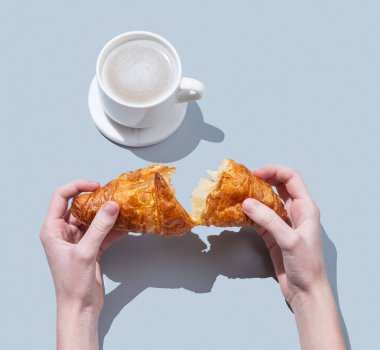 Mavi arka planda bir fincan kapuçino ya da latte kahveyle taze kruvasan kıran eller. Kafe reklamları ya da Fransız kahvaltı konsepti için yaratıcı bir model. Üst görünüm ve kopyalama alanı.