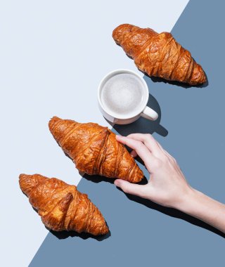 Mavi arka planda taze kruvasan, bir fincan kapuçino ya da pastalı ve gölgeli latte kahve. Kafe reklamları ya da Fransız kahvaltı konsepti için yaratıcı bir model. Üst görünüm ve kopyalama alanı