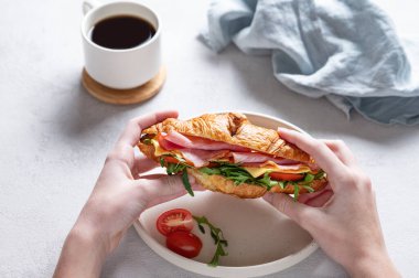 Elleri taze kruvasan sandviçi, jambon, peynir, domates ve roka ile hafif bir arka planda bir fincan kahve ve peçeteyle birlikte. Sağlıklı bir kahvaltı ya da brunch konsepti. Üst görünüm.