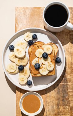 Tahtada muz ve yaban mersinli fıstık ezmeli tost ve bir fincan taze kahve. Amerikan kahvaltısı için tatlı diyet ve sağlıklı atıştırmalıklar kavramı. Üst görünüm ve kopyalama alanı.