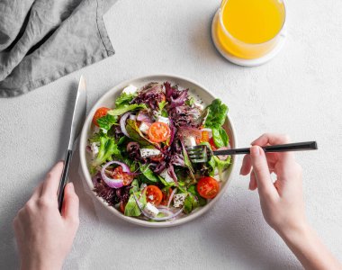 Elleri çatallı ve bıçaklı sebze salatası (Yunan salatası), domates, peynir, marul, zeytin ve soğanlı hafif zemin üzerinde bir bardak meyve suyu ve peçeteyle. Sağlıklı beslenme konsepti.