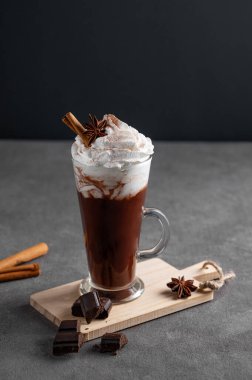 Krem şantili sıcak çikolata, tarçın ve koyu arkaplanda uzun bir bardakta baharatlar. Cafe reklamları veya afişler için tatlı içecek konsepti. Boşluğu kopyala.