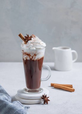 Krem şantili sıcak çikolata, tarçın ve baharatlar arka planda hafif bir sürahi ile uzun bir bardakta. Cafe reklamları veya afişler için tatlı içecek konsepti. Boşluğu kopyala.