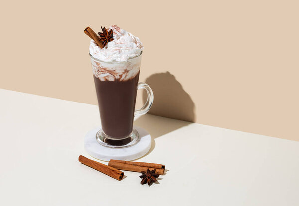 Hot chocolate with whipped cream, cinnamon and spices in a tall glass on a white and beige background with shadow. Sweet drink concept for cafe menu advertising or banner. Copy space.