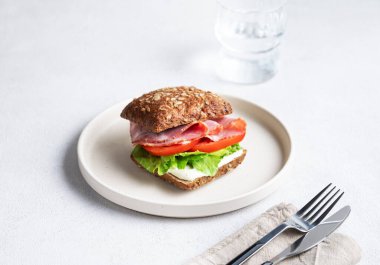 Jambonlu, peynirli, domatesli, marullu ve soslu tahıllı sandviç hafif bir zemin üzerinde bir bardak suyla. Sağlıklı ve diyet kahvaltı konsepti. Boşluğu kopyala.