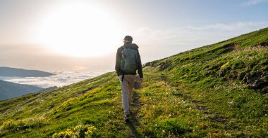 Sırt çantalı bir turist, gün doğumunda ya da gün batımında arkasında bulutlar olan izleyiciye sırtını dönük olarak yeşil bir dağ yolunda yürüyor. Doğa yolculuğu konsepti.