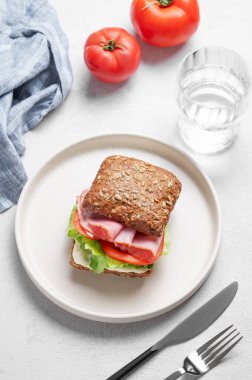 Prosciutto, peynir, domates, marul ve sostan yapılmış tahıllı sandviç hafif bir arka planda bir tabakta, bir bardak su, peçete ve çatalla. Sağlıklı ve diyet kahvaltı kavramı. 