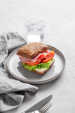 Prosciutto, peynir, domates, marul ve soslu tahıllı sandviç hafif bir arka planda bir tabakta, bir bardak su ve sabah ışığıyla birlikte. Sağlıklı ve diyet kahvaltı kavramı. 