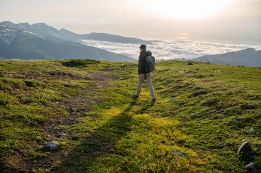 Sırt çantalı bir turist, güneşin doğuşunda veya batışında yeşil bir dağ yolu boyunca yürür. Altında bulutlar ve arka planında karlı tepeler vardır. Doğa yolculuğu konsepti.