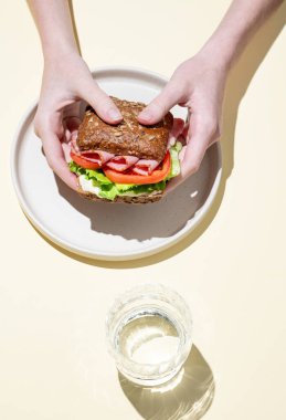 Jambonlu, marullu, domatesli ve soslu taze bir sandviçi pastel sarısı arka planda bir tabakta, bir bardak suyla tam tahıllı ekmeğin üzerinde tutan eller. Sağlıklı beslenme kavramı. Üst görünüm, alanı kopyala