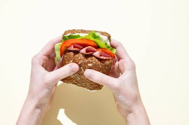 Jambonlu, marullu, domatesli ve soslu taze bir sandviçi pastel sarısı arka planda tam tahıllı ekmeğin üstünde tutan eller. Sağlıklı beslenme kavramı. Üst görünüm, alanı kopyala.