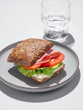 Jambonlu tahıllı sandviç, domates, marul ve sos gri zemin üzerinde bir bardak suyla. Kafe reklamları veya afişler için sağlıklı kahvaltı konsepti. Boşluğu kopyala.