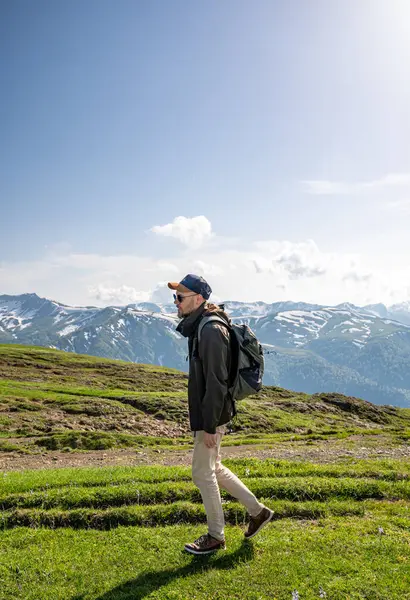 Sırt çantalı bir turist güneşli bir günde yeşil bir dağ yolunda yürüyor, aşağısı bulutlu ve arka planında karlı zirveler var. Doğa yolculuğu konsepti.