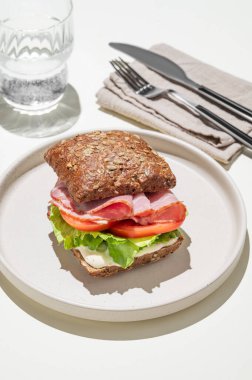 Jambonlu tahıllı sandviç, domates, marul ve sosla hafif arka planda bir bardak suyla. ve çatal. Kafe menüsü reklamcılığı ya da afişi için sağlıklı kahvaltı kavramı. 