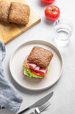 Prosciutto, peynir, domates, marul ve sostan yapılmış tahıllı sandviç hafif bir arka planda bir tabakta, bir bardak su, peçete ve çatalla. Sağlıklı ve diyet kahvaltı kavramı. 