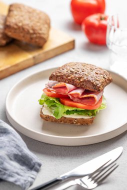 Prosciutto, peynir, domates, marul ve sostan yapılmış tahıllı sandviç hafif bir arka planda bir tabakta, bir bardak su, peçete ve çatalla. Sağlıklı ve diyet kahvaltı kavramı. 