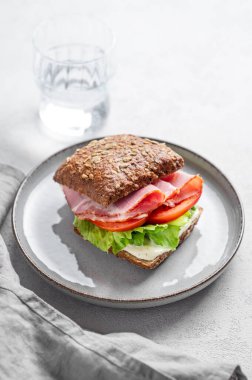 Prosciutto, peynir, domates, marul ve soslu tahıllı sandviç hafif bir arka planda bir tabakta, bir bardak su ve sabah ışığıyla birlikte. Sağlıklı ve diyet kahvaltı kavramı. 