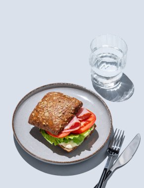 Prosciutto, domates, marul ve sostan yapılmış tahıllı sandviçle mavi zemin üzerinde bir bardak su. Kafe reklamları veya afişler için sağlıklı kahvaltı konsepti. Üst görünüm.