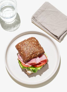 Jambonlu tahıllı sandviç, domates, marul ve sosla hafif arka planda bir bardak suyla. Kafe reklamları veya afişler için sağlıklı kahvaltı konsepti. Üst görünüm.