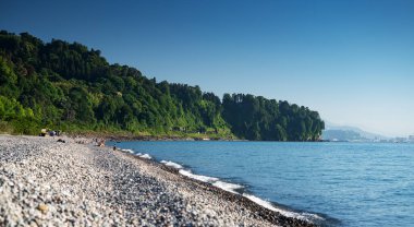 Güneşli bir günde, yeşil ormanın yanında dinlenen insanların, sakin mavi denizin ve uzak dağların olduğu güneşli bir sahilin panoramik manzarası. Batumi, Georgia.