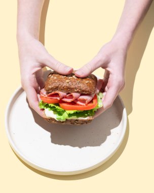 Ellerinde jambon, marul, domates ve sosla taze bir sandviçi sarı bir arka planda tam tahıllı ekmeğin üzerinde tutuyorlar. Sağlıklı beslenme kavramı. Üst görünüm, alanı kopyala.