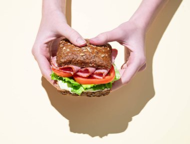 Jambonlu, marullu, domatesli ve soslu taze bir sandviçi pastel sarısı arka planda tam tahıllı ekmeğin üstünde tutan eller. Sağlıklı beslenme kavramı. Üst görünüm, alanı kopyala.